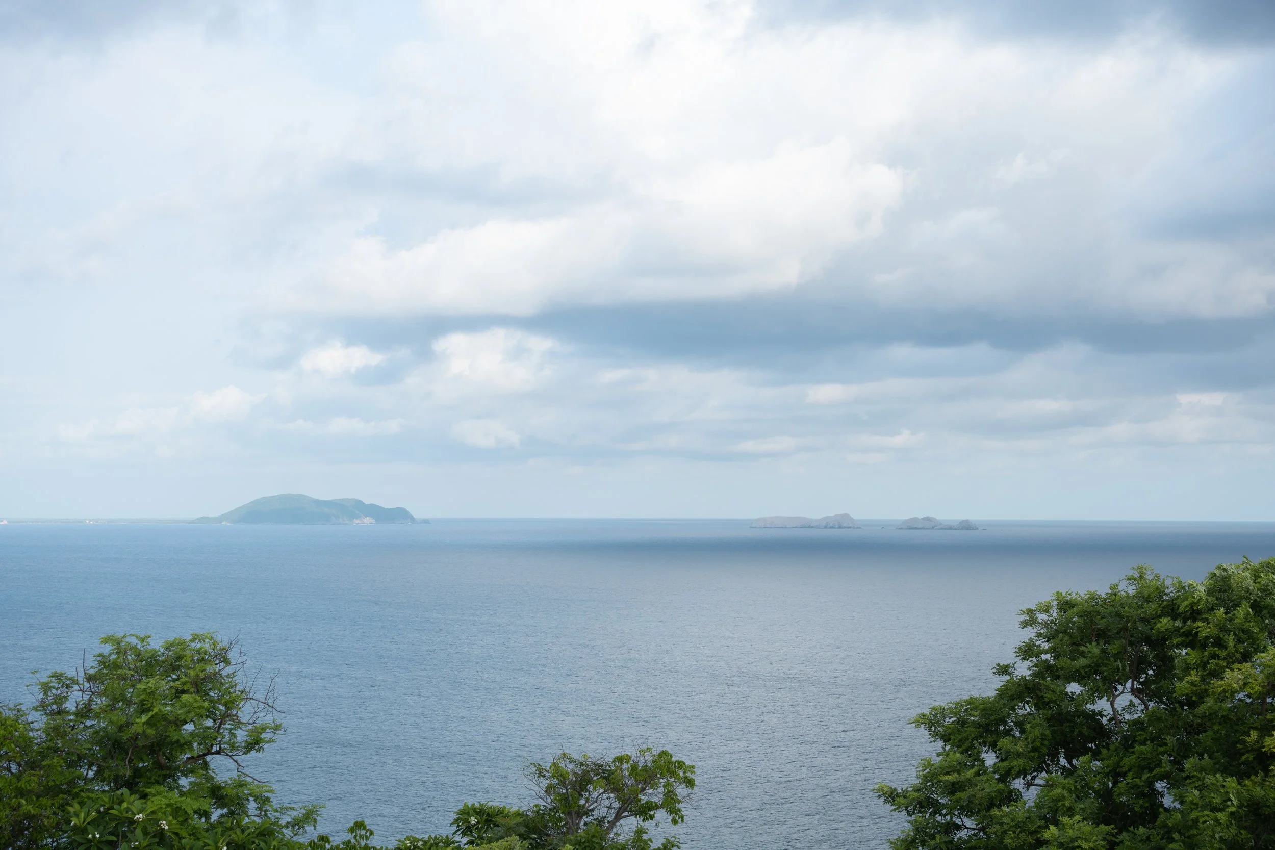 Vista panorámica de un mar con pequeñas islas en el horizonte, nubes grises en el cielo y árboles verdes en primer plano.