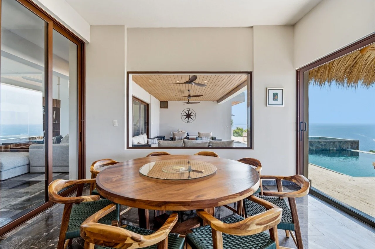 Comedor con vista al mar y sala de estar al fondo, con muebles de madera y paredes blancas.