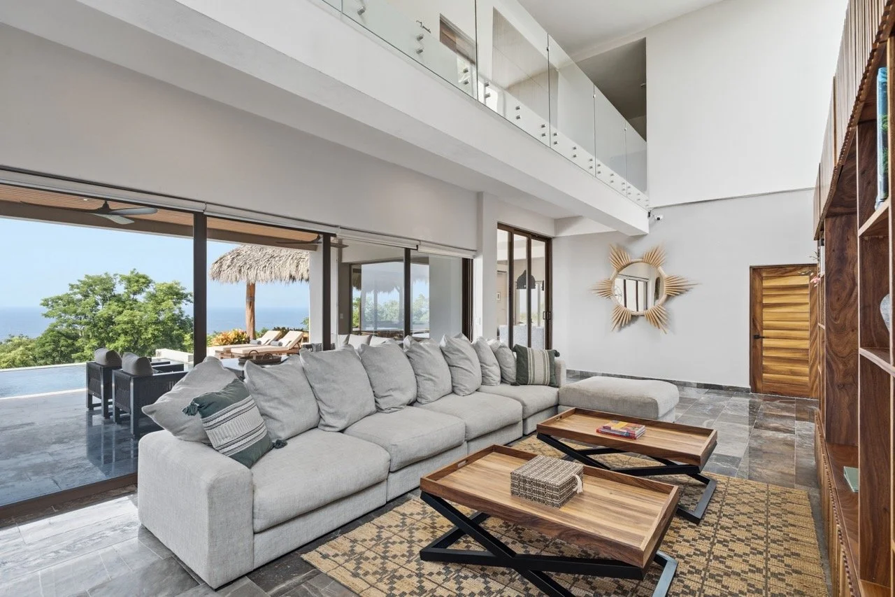 Sala moderna con vista al exterior, sofá gris, mesas de madera y decoración en tonos neutrales.