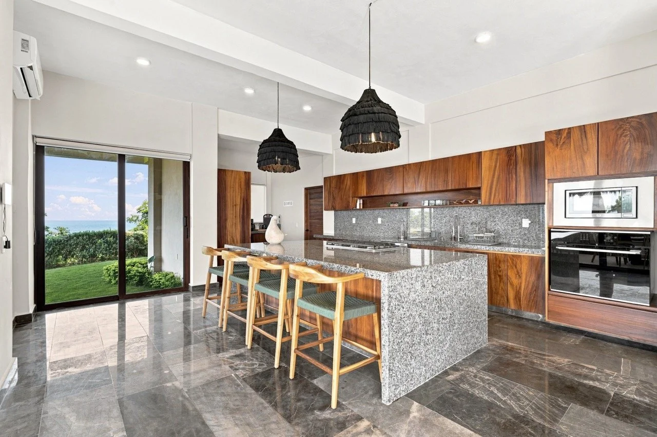 Cocina moderna con encimeras de granito y gabinetes de madera, barra con cuatro sillas y vista hacia el jardín y el mar a través de una puerta corrediza de cristal.