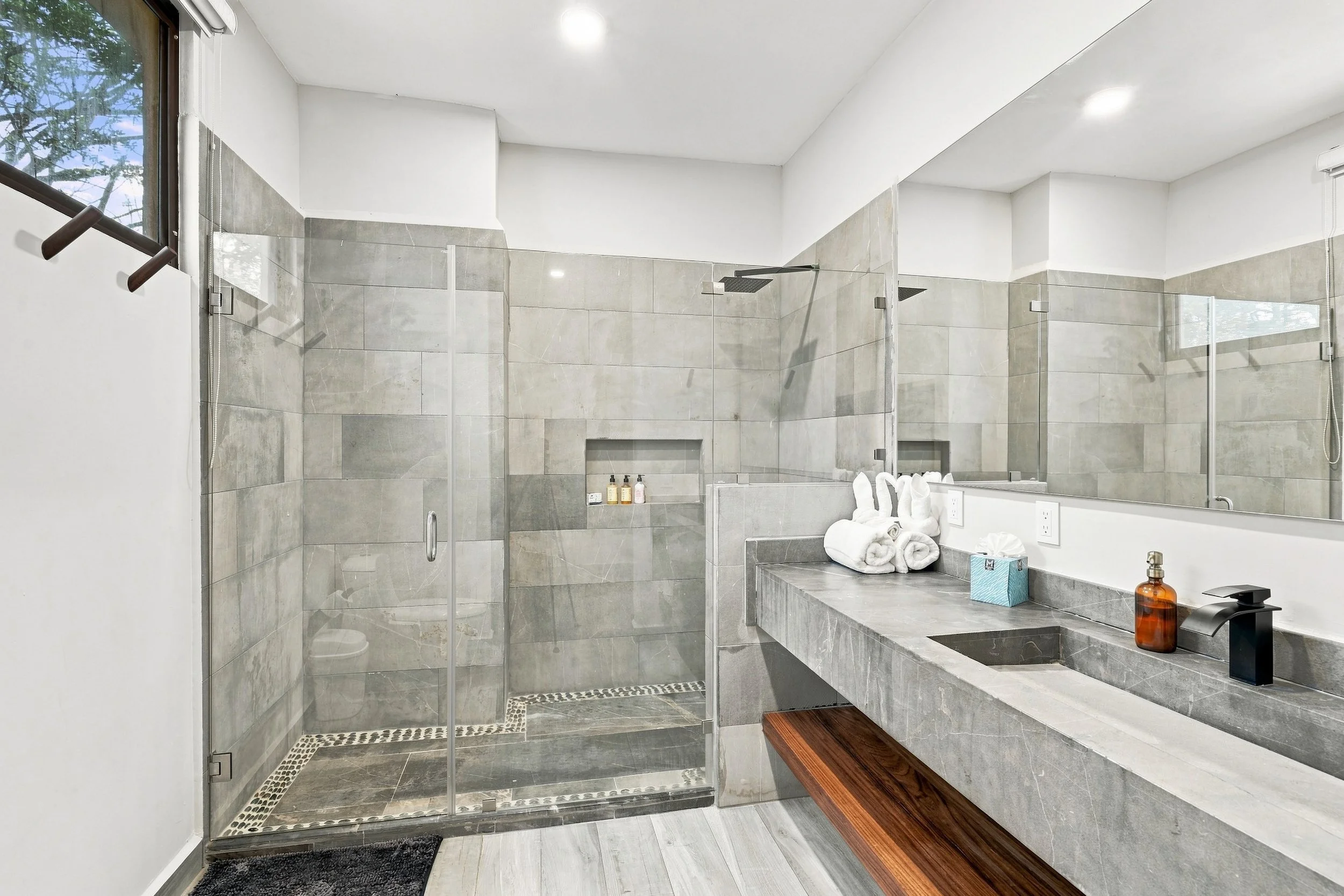 Baño moderno con ducha de cristal, lavabo de piedra, espejo grande y accesorios de cuidado personal, en tonos gris y blanco.