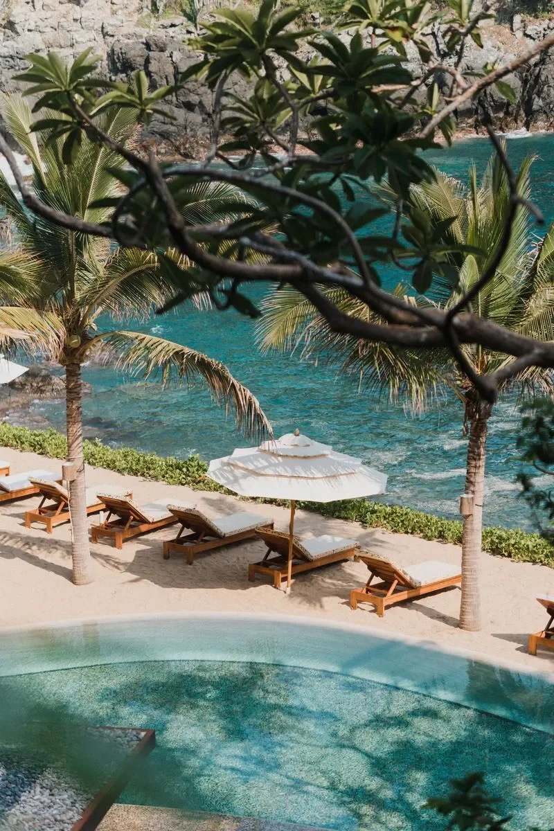 Vista de una playa tropical con hamacas de madera y sombrillas blancas, palmeras y agua cristalina, vista parcialmente bloqueada por ramas y hojas de árboles.
