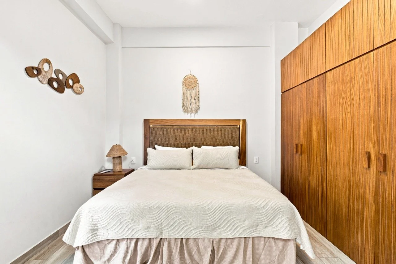 Habitación con cama, cabecera de mimbre, pared blanca, armario de madera, lámpara de noche de madera y decoraciones de madera en la pared.