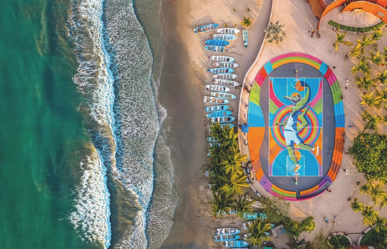 Foto aérea de una playa con olas en el mar, zonas de arena, palmeras y una cancha de baloncesto decorada con arte colorido.