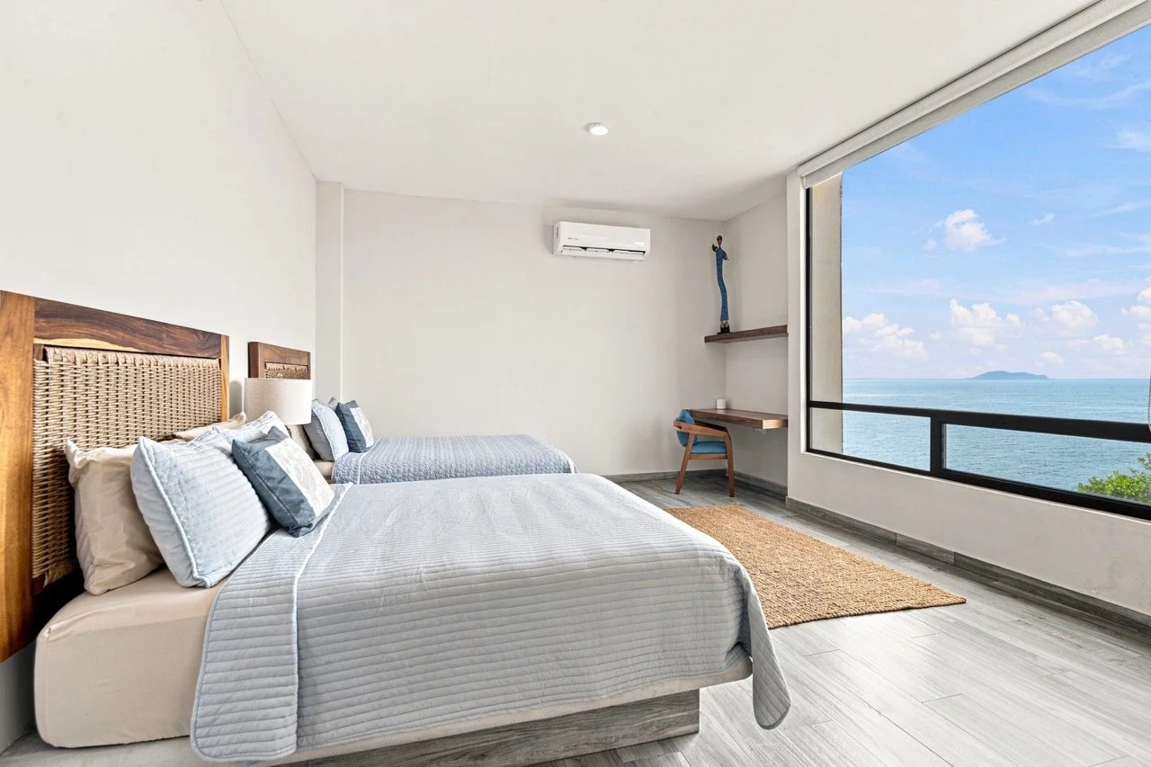 Habitación moderna con vista al mar, dos camas con ropa de cama blanca y cojines azules, suelo de madera, y una gran ventana con vista al océano y cielo azul.