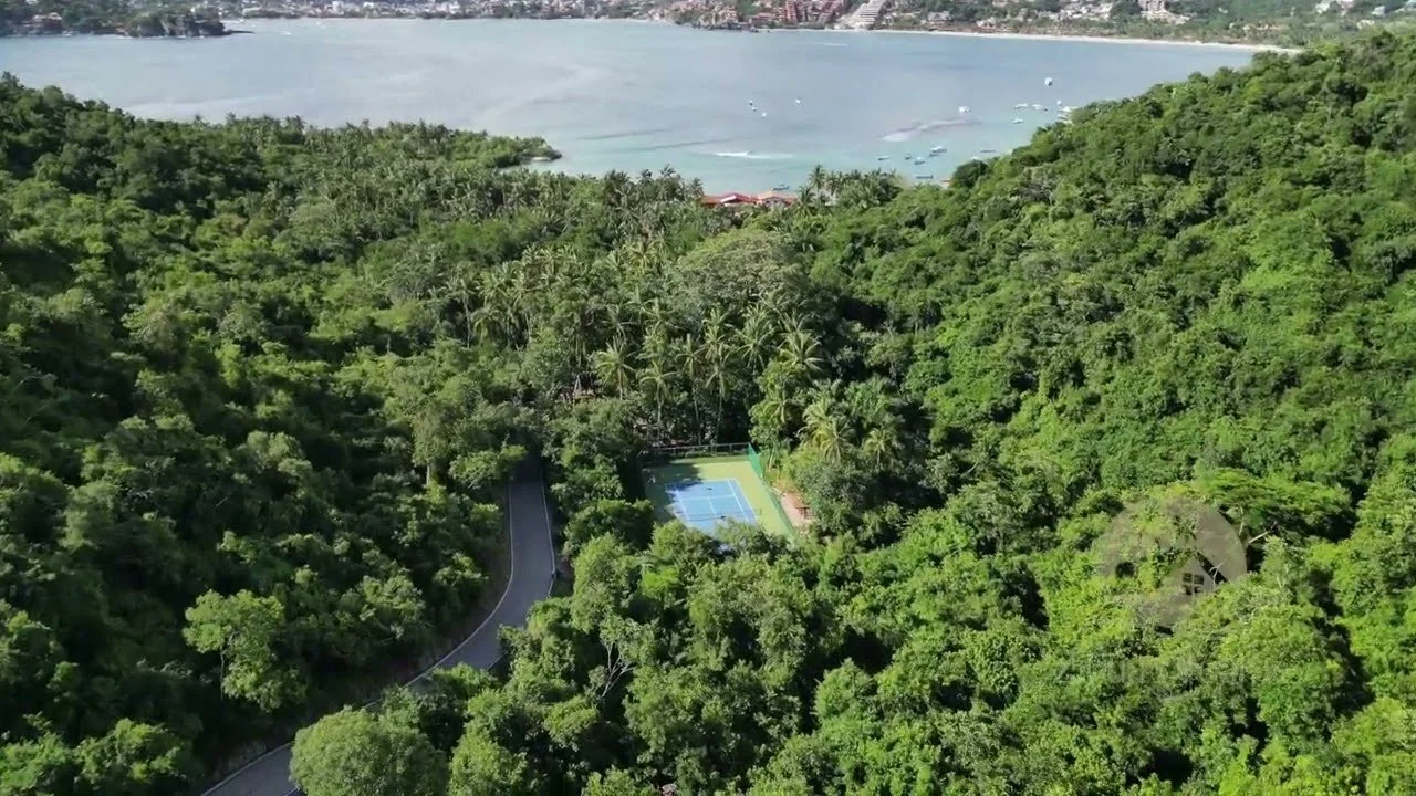 Vista aérea de una zona boscosa con una cancha de tenis y un camino, cerca de la costa con agua y barcos pequeños en el fondo.