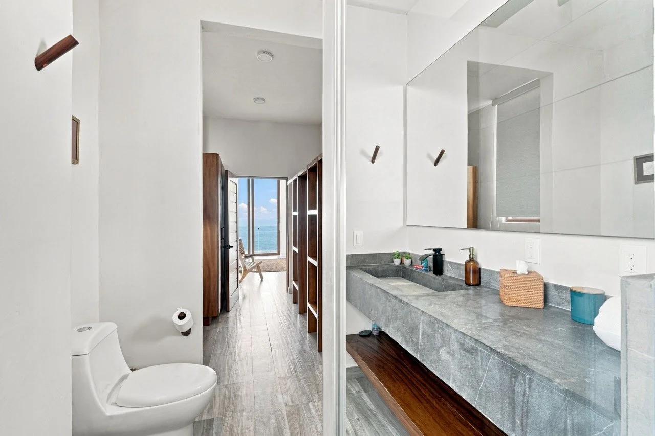 Vista de un baño con lavabo de piedra gris, espejo grande y accesorios de tocador, y una sala con vista al mar por una ventana grande