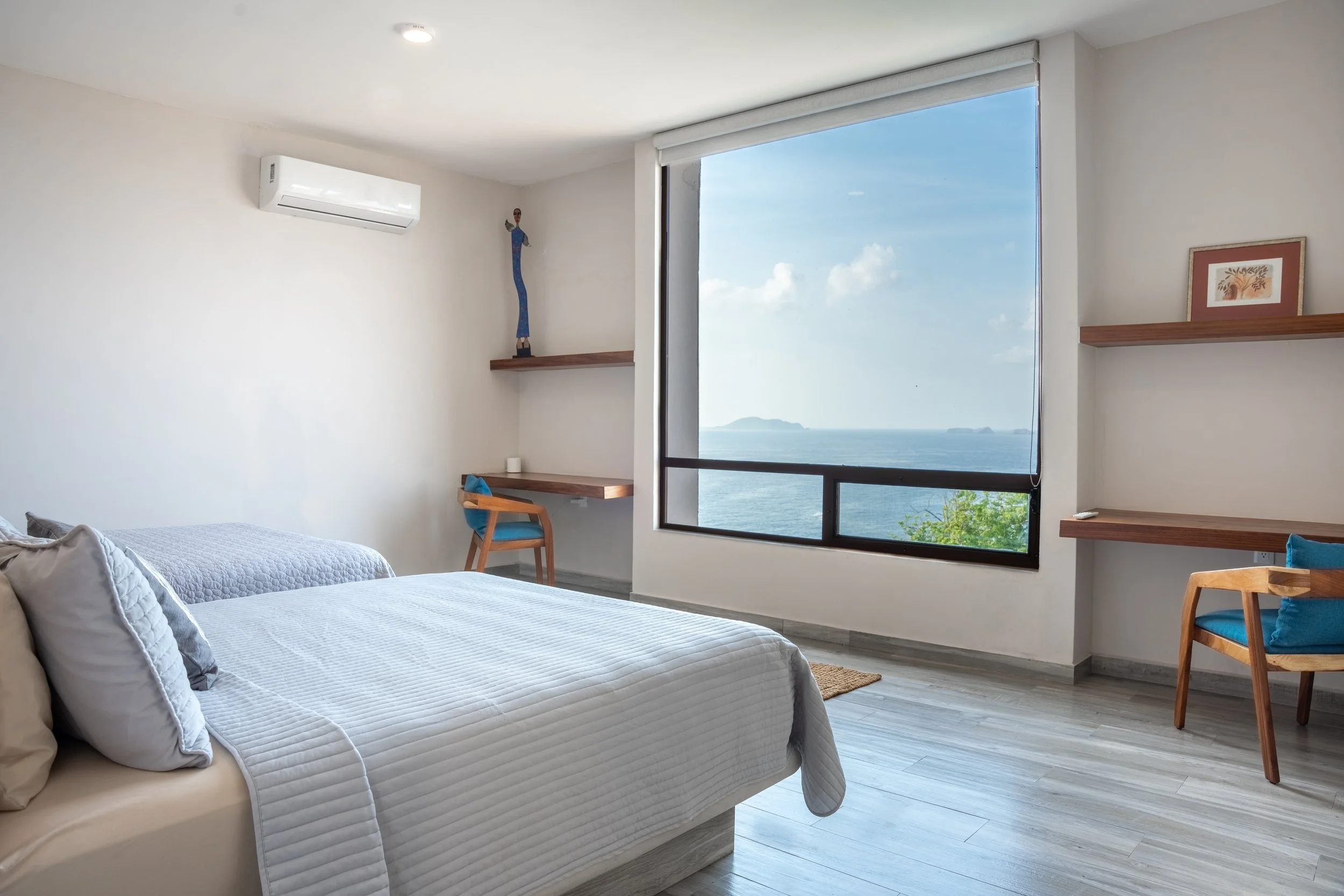 Habitación moderna con cama, vista al mar, estanterías de madera y una silla de madera con cojín azul.