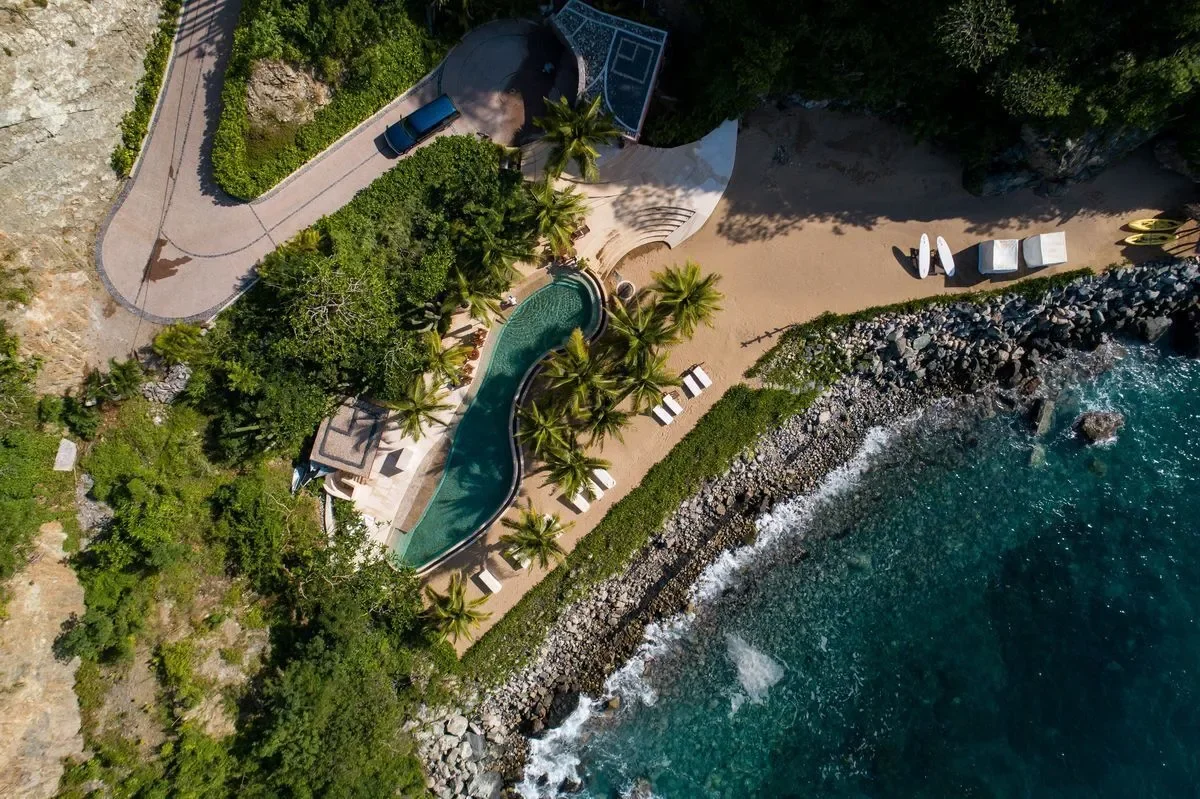 Vista aérea de una piscina con forma curva en una zona junto al mar, rodeada de palmeras y áreas de descanso con tumbonas y sombrillas, en un entorno tropical con vegetación y rocas en la orilla del océano.