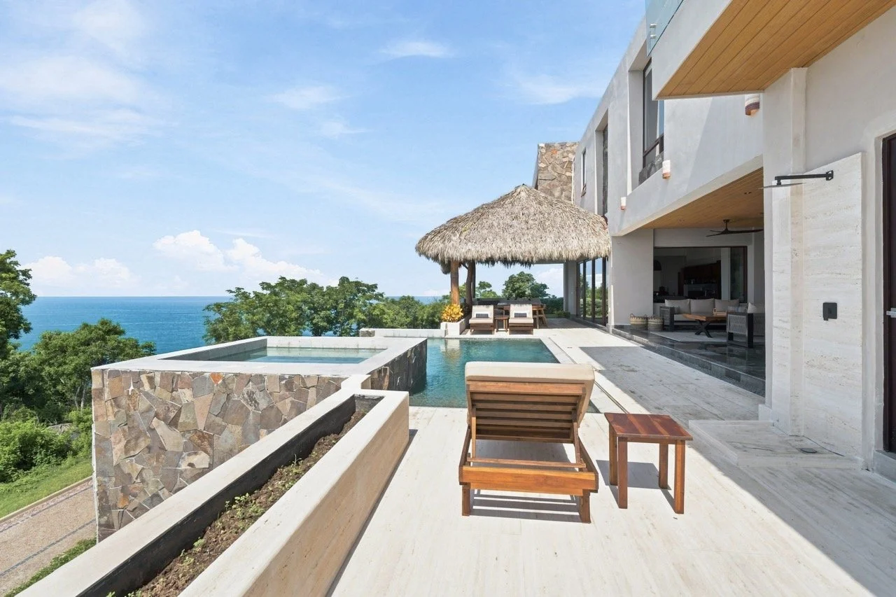 Vista de una terraza moderna con piscina, tumbonas de madera, sombrilla de paja y vista al océano, parte de una casa con paredes de piedra y áreas abiertas.