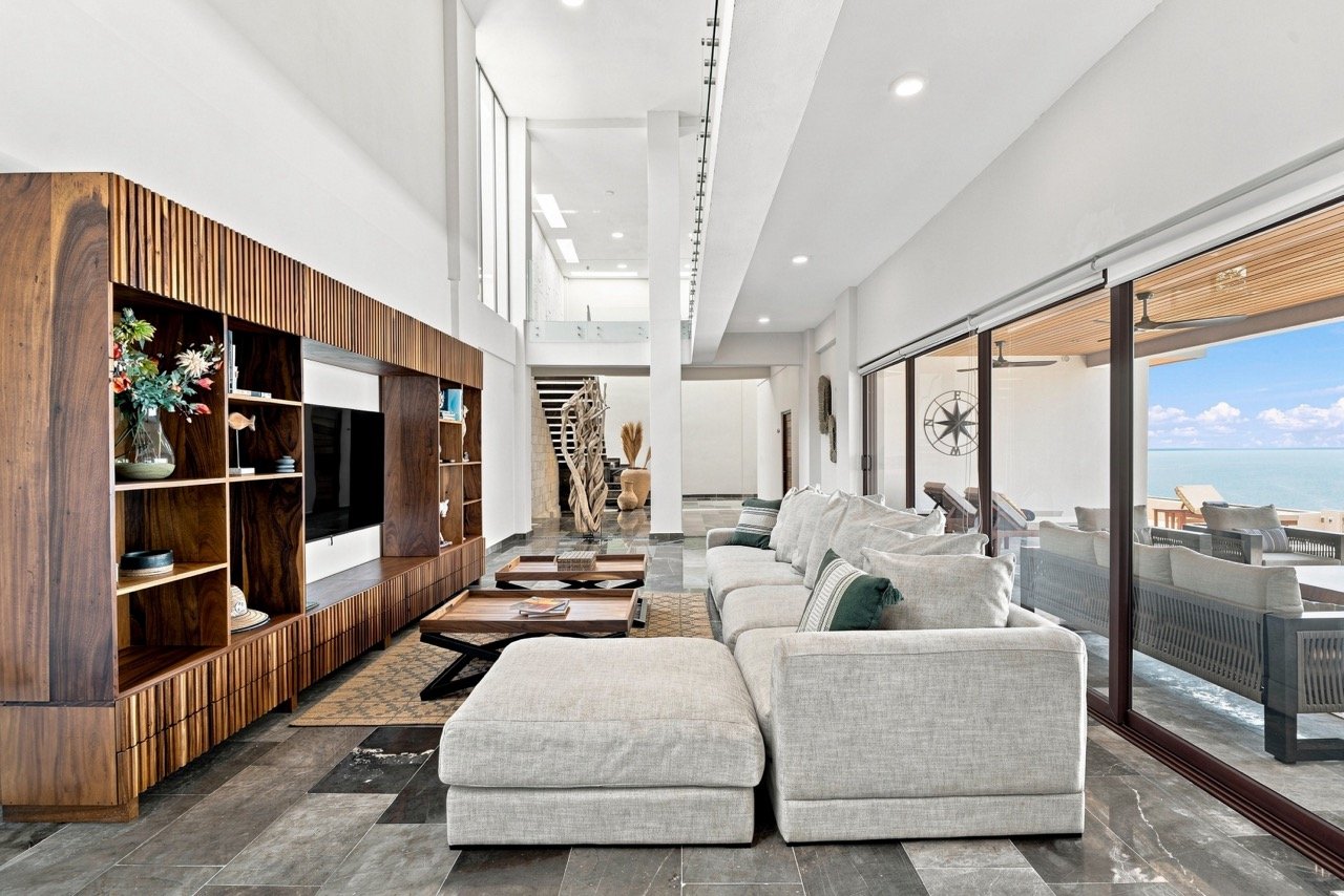 Sala de estar moderna con sillón de color beige, estantería de madera, televisión, decoración y vista al mar a través de ventanas grandes