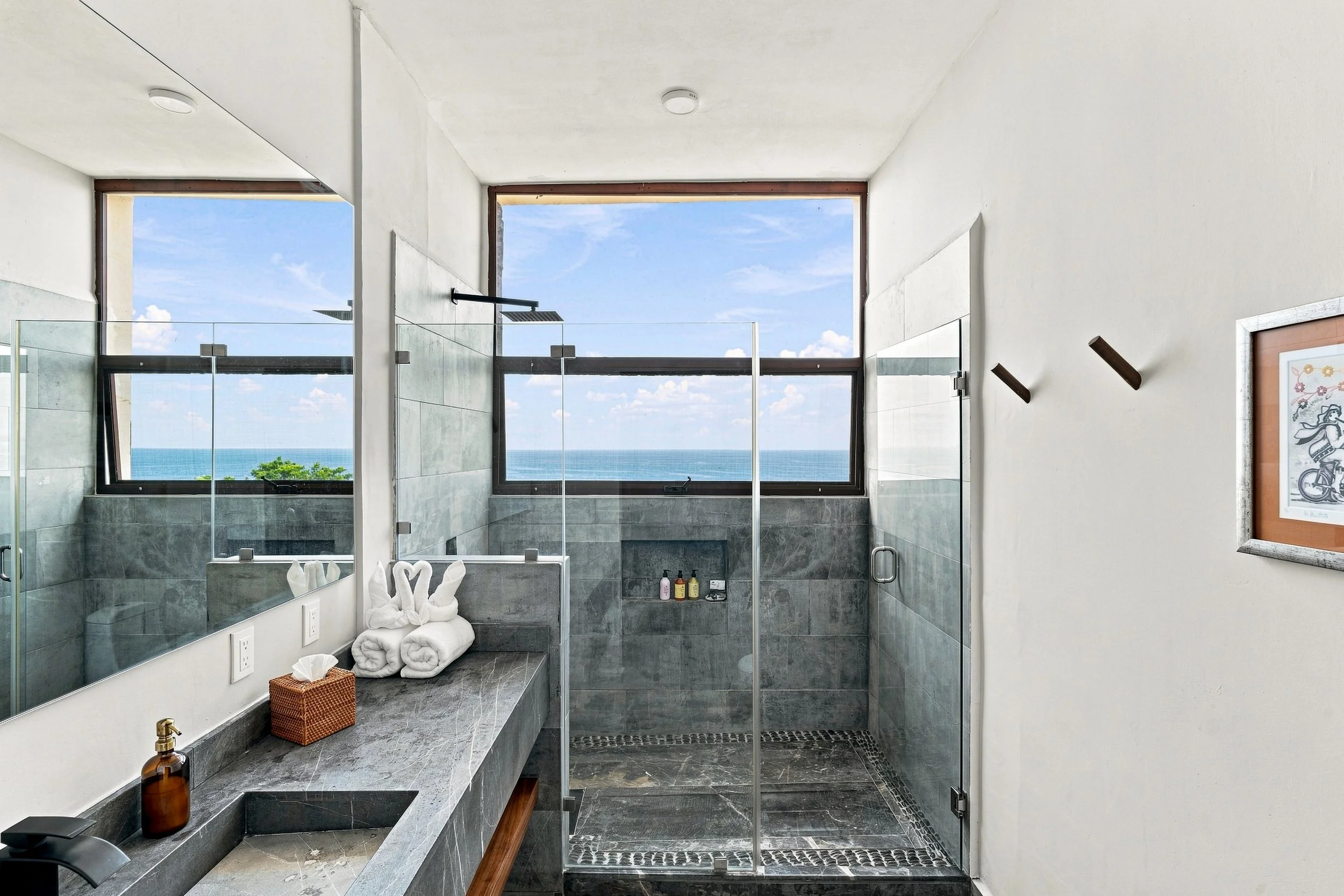 Baño con vista al mar a través de ventanas grandes, ducha con puertas de vidrio, lavabo con espejo y toallas dobladas en forma de cisne.