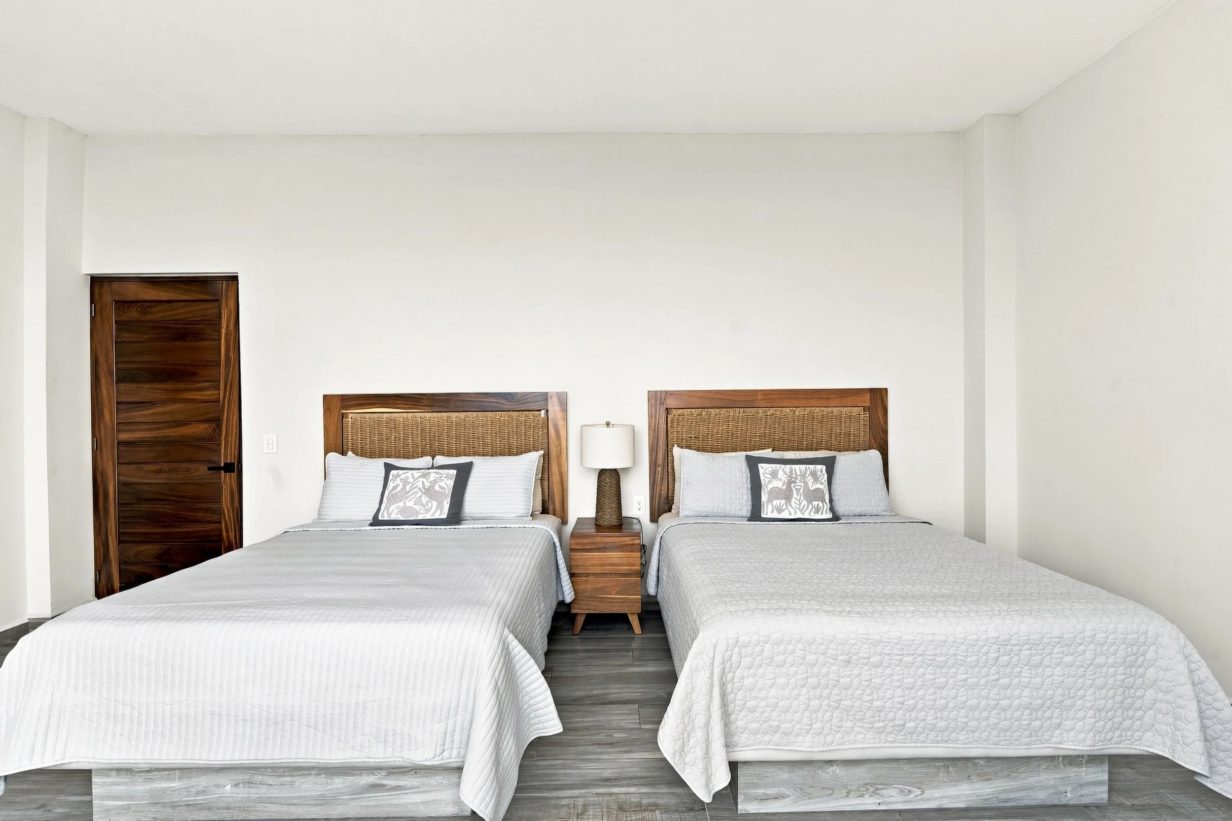 Habitación con dos camas de matrimonio, cabeceras de madera, lámpara entre ellas, y paredes blancas.