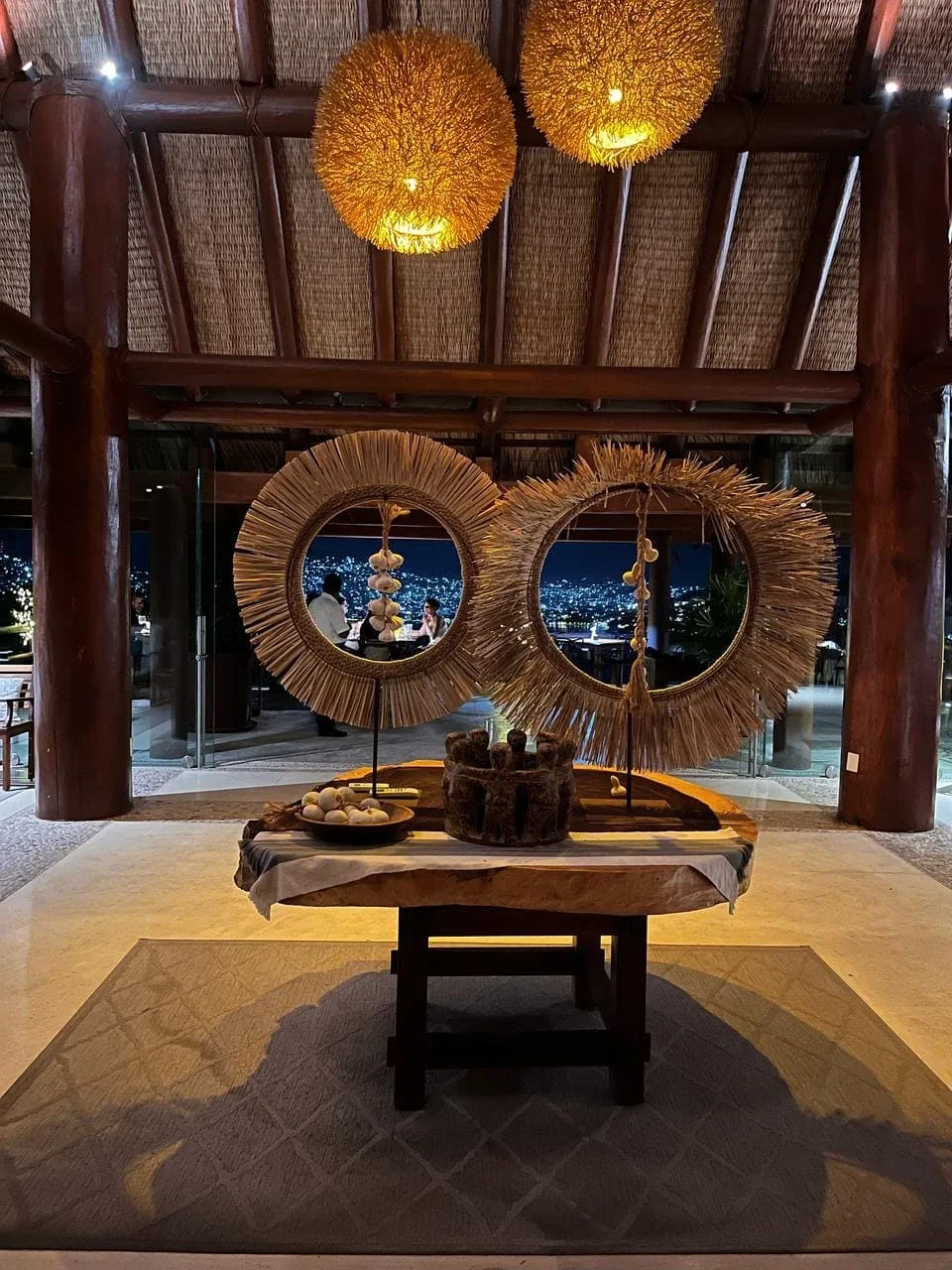 Decoración rústica con luces cálidas, todo en un espacio de madera, con espejo de corona de paja, mesa con objetos decorativos, vista nocturna al fondo con ciudad y personas en un ambiente acogedor.