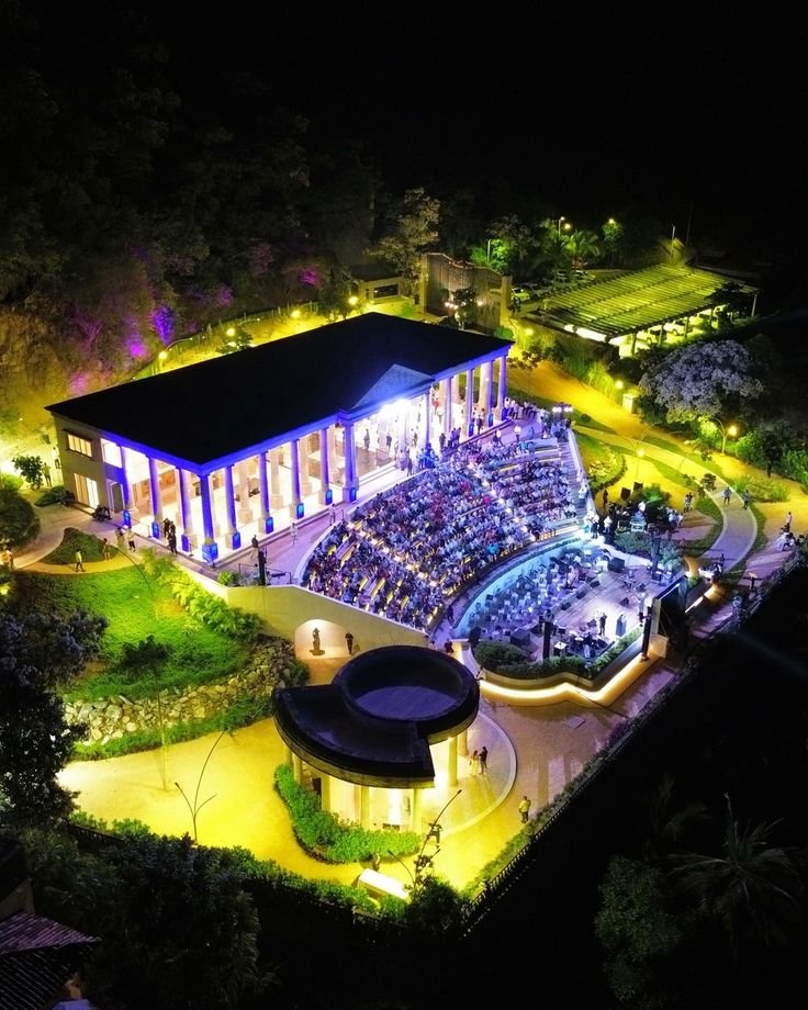 Vista aérea de un anfiteatro al aire libre iluminado de noche, lleno de gente, con un edificio moderno con columnas frente a una zona de árboles y jardines.