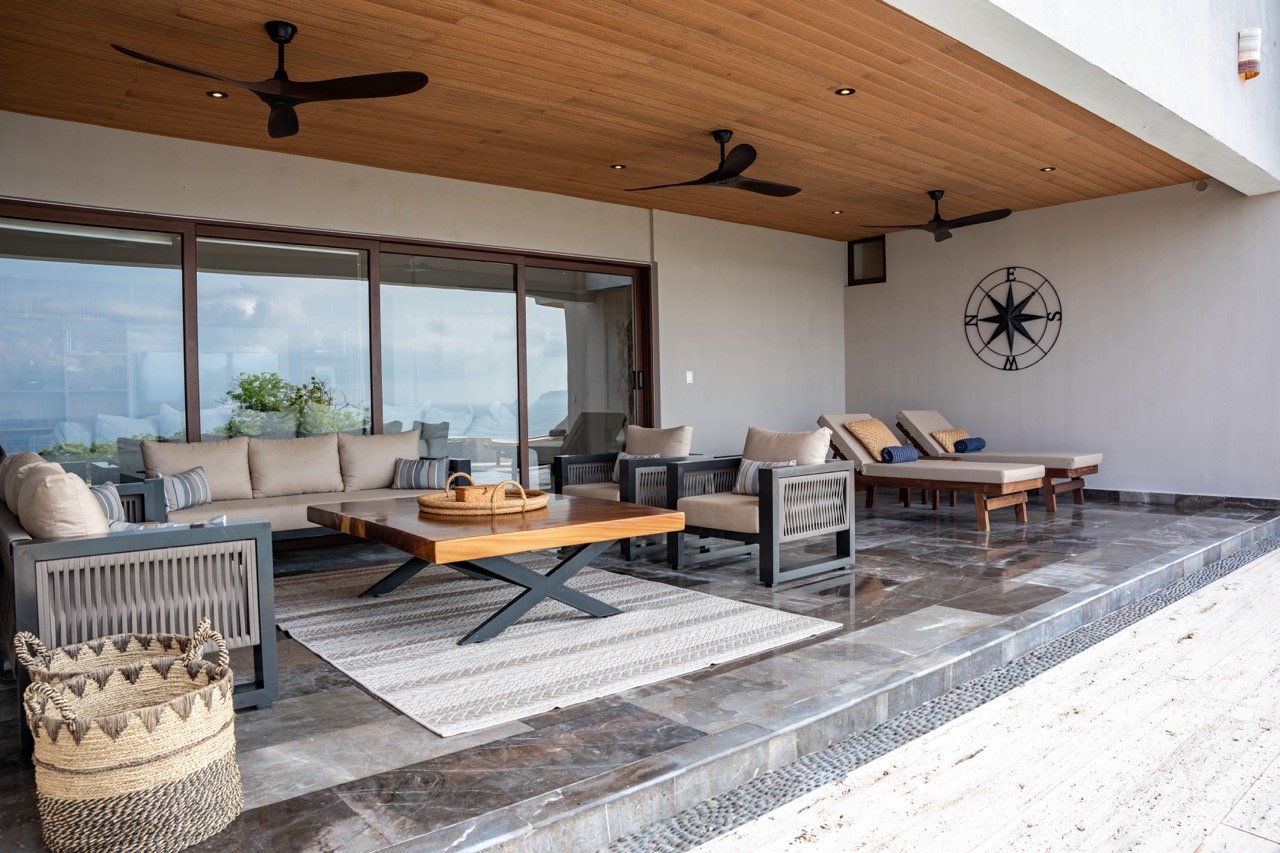 Terraza de una casa con muebles de exterior, sillones, mesas, cojines y tumbonas, con vista al mar y un cielo nublado.