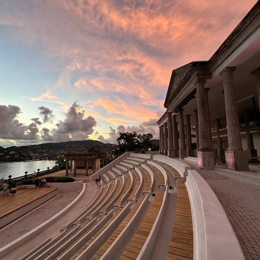 Escenario al atardecer con un edificio de columnas clásicas, un río y colinas en el fondo, y un espacio con asientos en primer plano.