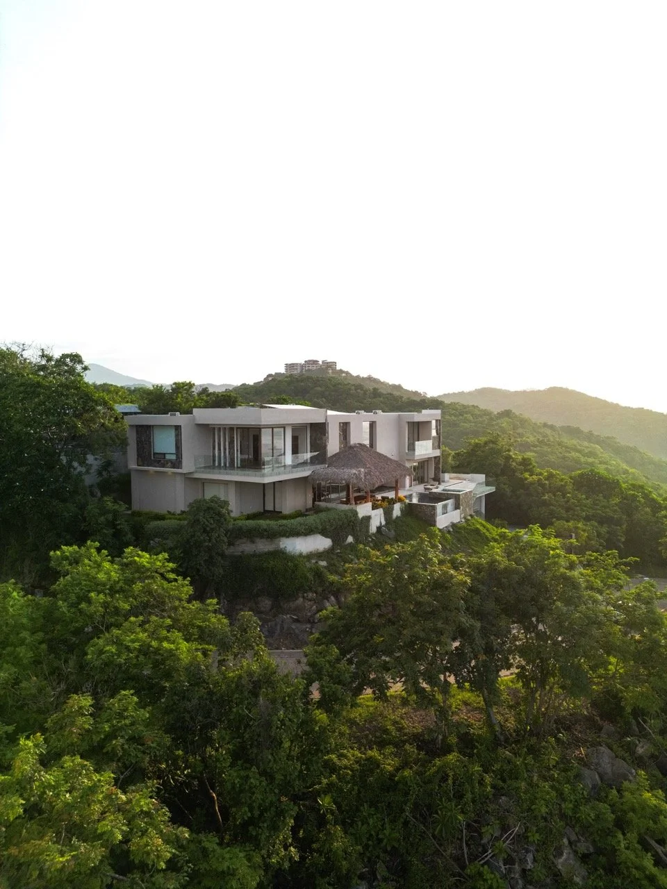 Casa moderna en una colina rodeada de árboles verdes con montañas al fondo.