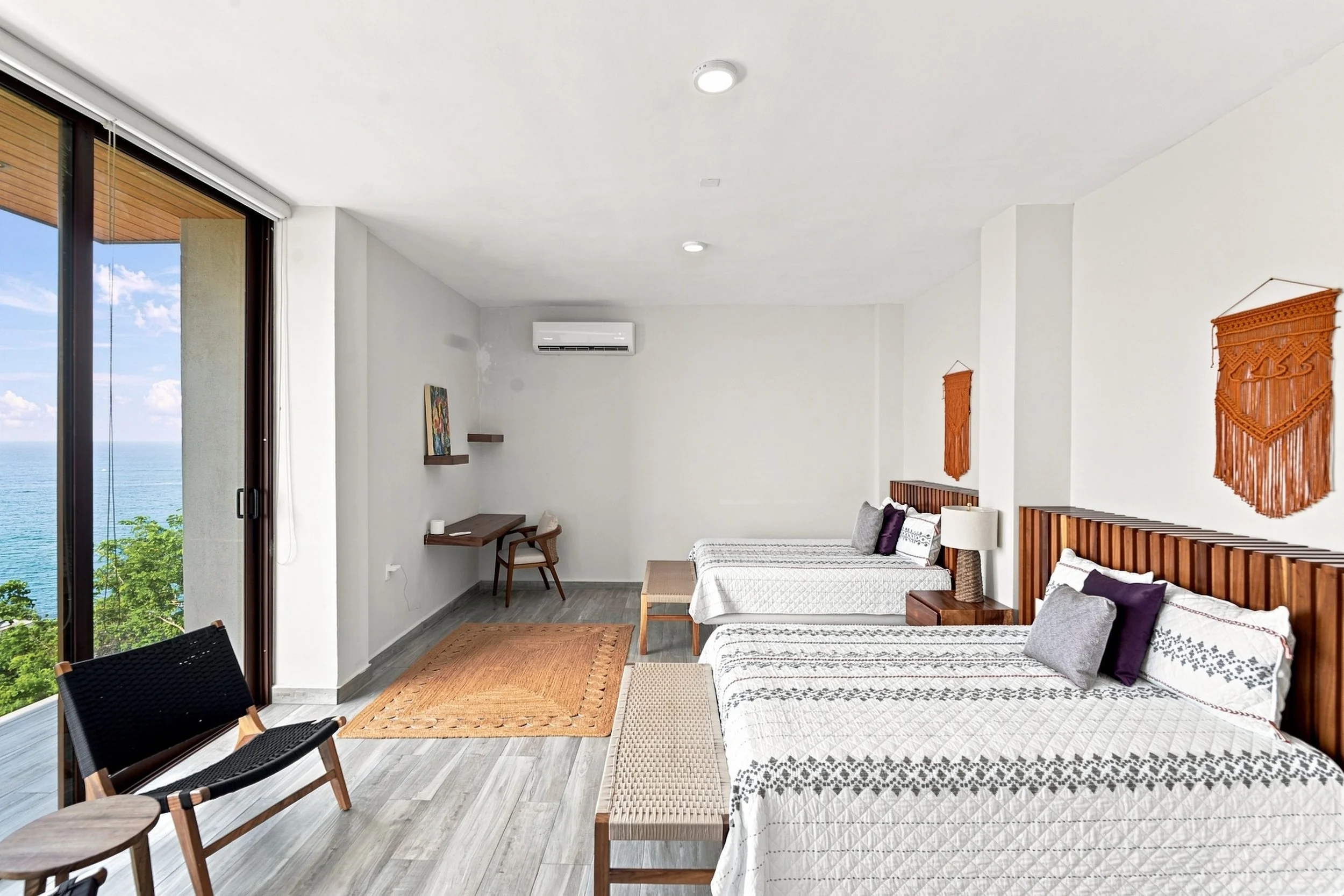 Habitación con vista al mar, dos camas con ropa de cama blanca y detalles en morado y gris, paredes blancas, decoraciones de tapices de hilo en las paredes, ventanales grandes con vista al océano, aire acondicionado, y muebles de madera.