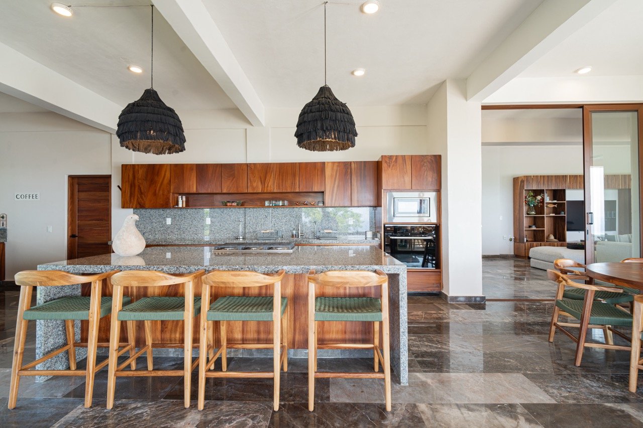 Cocina abierta con isla de granito, ocho sillas de madera con cojines verdes, armarios de madera, televisor y librería en el fondo, suelo de piedra y luminarias negras colgantes.