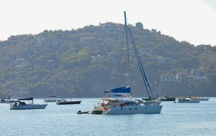 Yate de vela en el agua con otras embarcaciones y colina con edificios en el fondo.