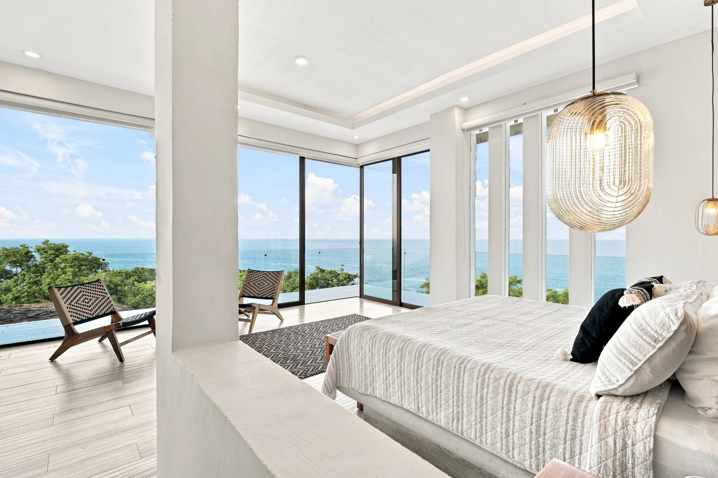 Habitación moderna con vista al mar, grandes ventanales, cama con colchas claras y cojines oscuros, lámpara colgante de diseño elegante, sillas de madera en el balcón, área luminosa y decorativa.