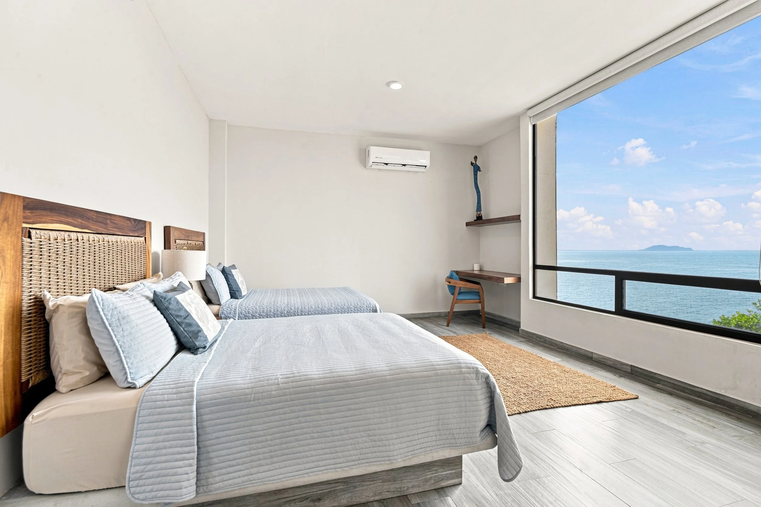 Habitación con vista al mar, dos camas con ropa de cama blanca y cojines, madera en cabecera y detalles, alfombra de yute y una ventana grande con vista al océano y cielo despejado.