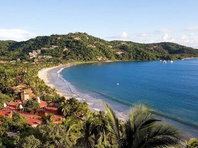 Vista de una playa con agua azul, árboles y casas en las colinas cercanas.