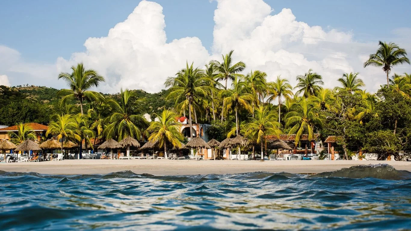 Vista de una playa tropical con palmeras, cabañas de paja y el mar en primer plano.