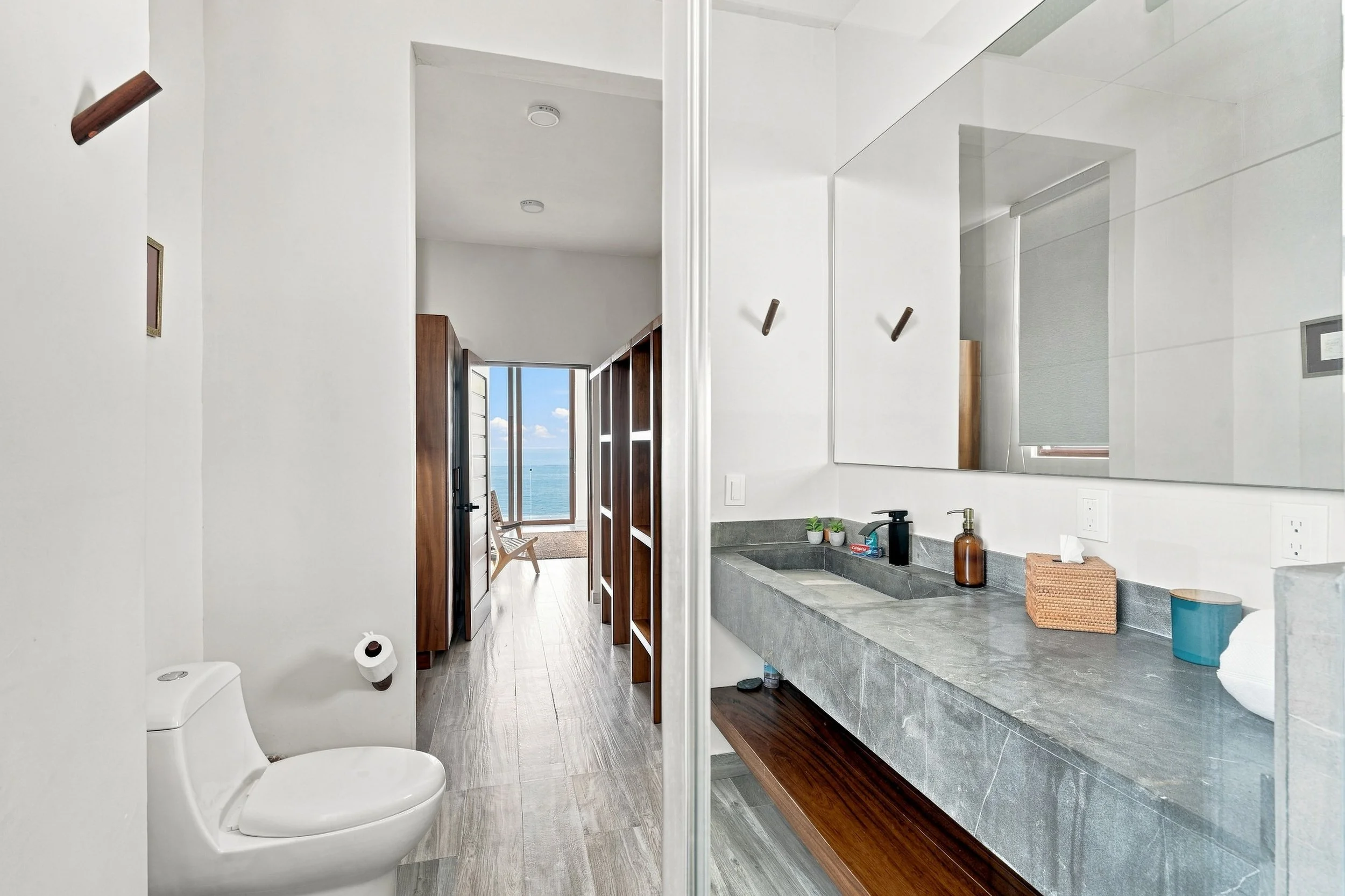 Baño con lavabo de piedra, espejo grande, objetos de higiene y un inodoro, con vista al exterior por la puerta abierta que muestra el mar y el cielo azul