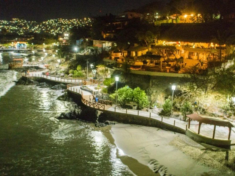 Camino peatonal iluminado cerca de la playa con olas rompiendo y casas iluminadas arriba de la colina, vista nocturna.