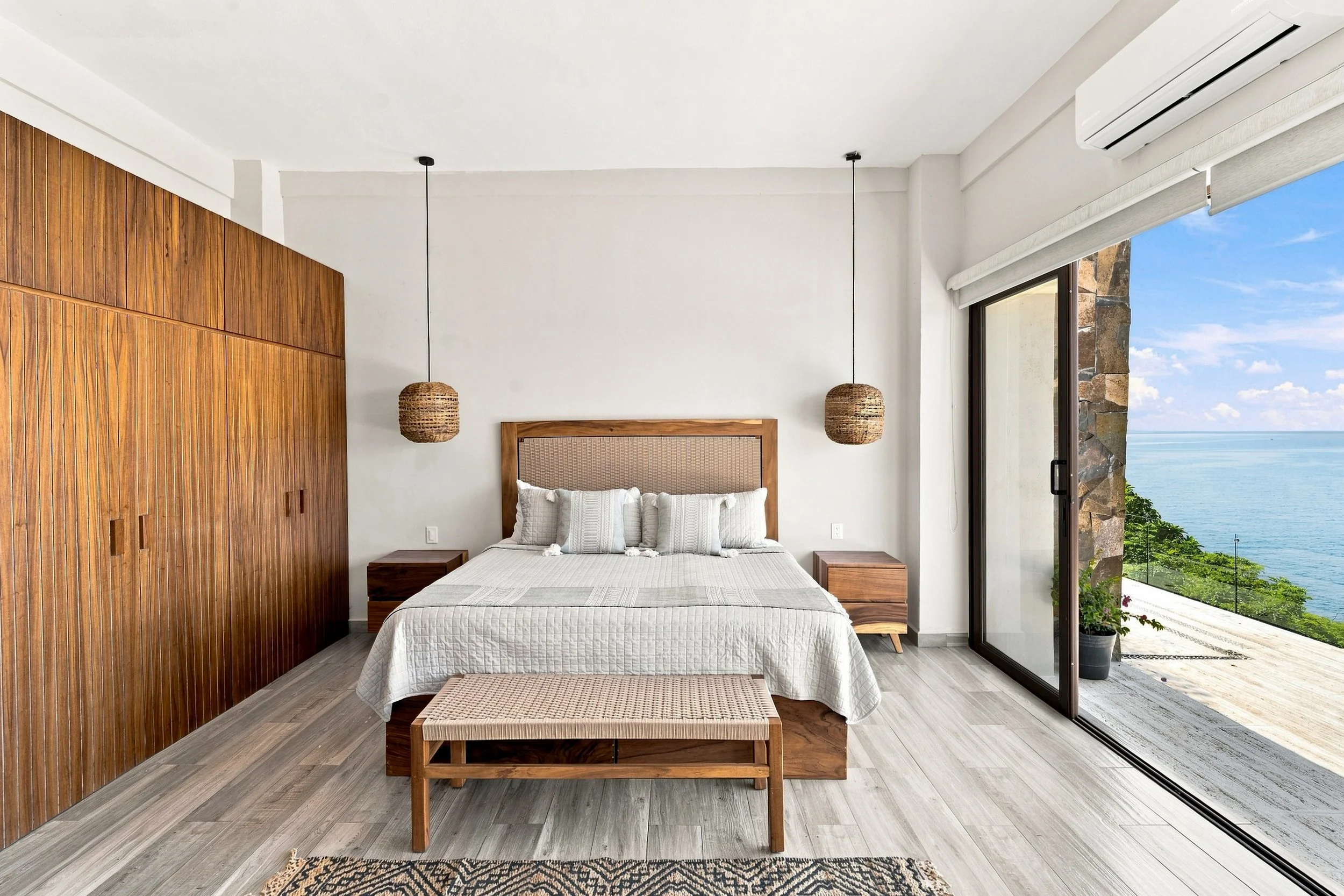 Habitación con cama de madera, cabecera de mimbre, dos mesitas de noche y lámparas de cuerda, vista al mar a través de un ventanal grande con piso de madera y paredes blancas, decoración moderna y natural.
