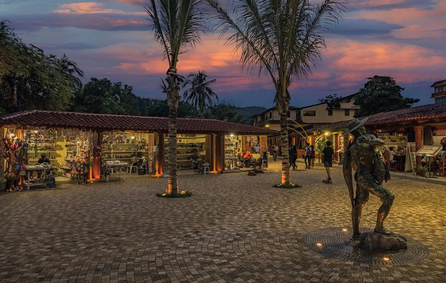 Plaza con puestos de artesanías, palmeras, tienda, esculturas y personas en el atardecer.