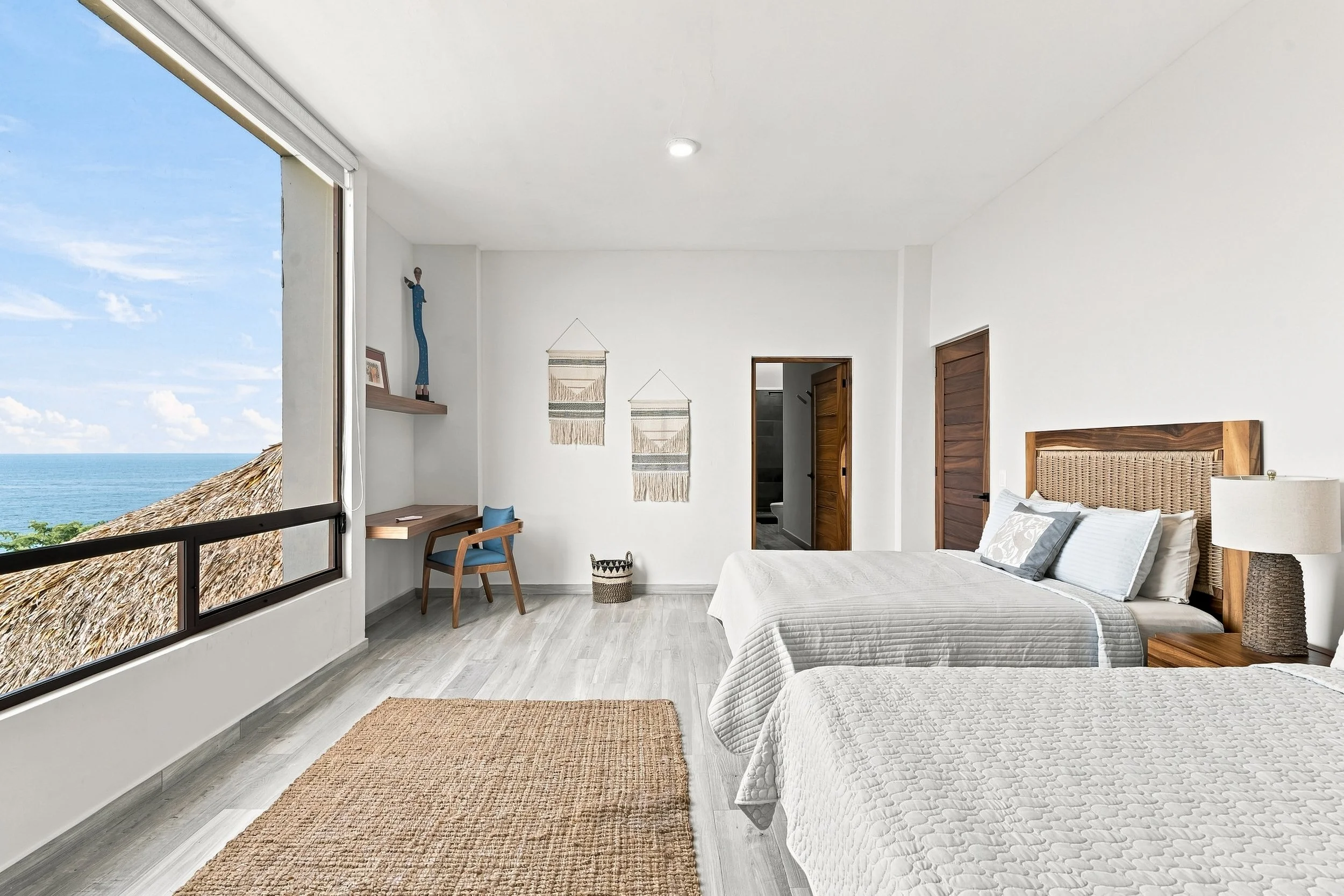 Habitación con vista al mar, tiene dos camas con cabecera de madera, paredes blancas, un espejo, sillón de madera con cojín azul, decoraciones en la pared y un tapete de yute, gran ventana con vista al mar y techo blanco con luz empotrada.