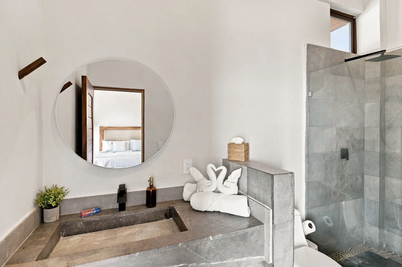 Baño moderno con lavabo de piedra, espejo redondo, decoración de toallas en forma de cisne, planta, y ducha con puertas de vidrio.