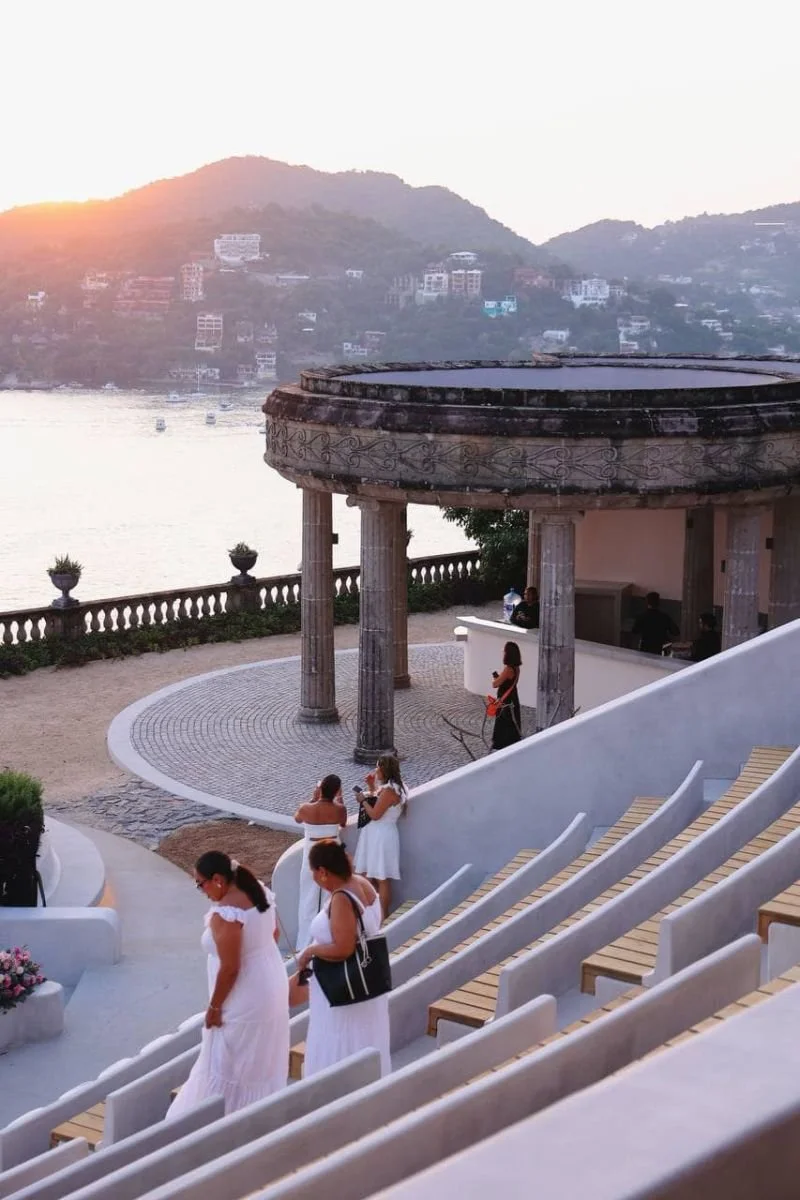 Un grupo de personas en una terraza con vista al río y a colinas, durante el atardecer, algunas vestidas de blanco y conversando.