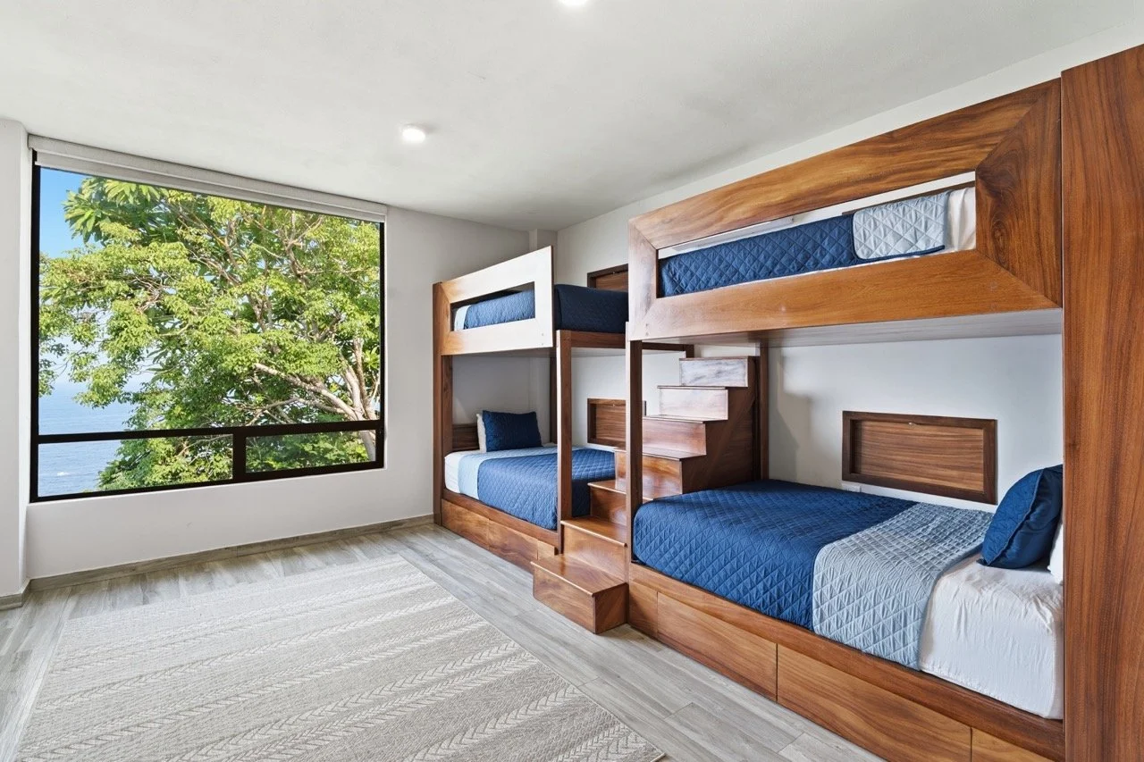 Habitación con camas literas de madera, ropa de cama azul y una ventana grande con vista a un árbol y el mar en el fondo.