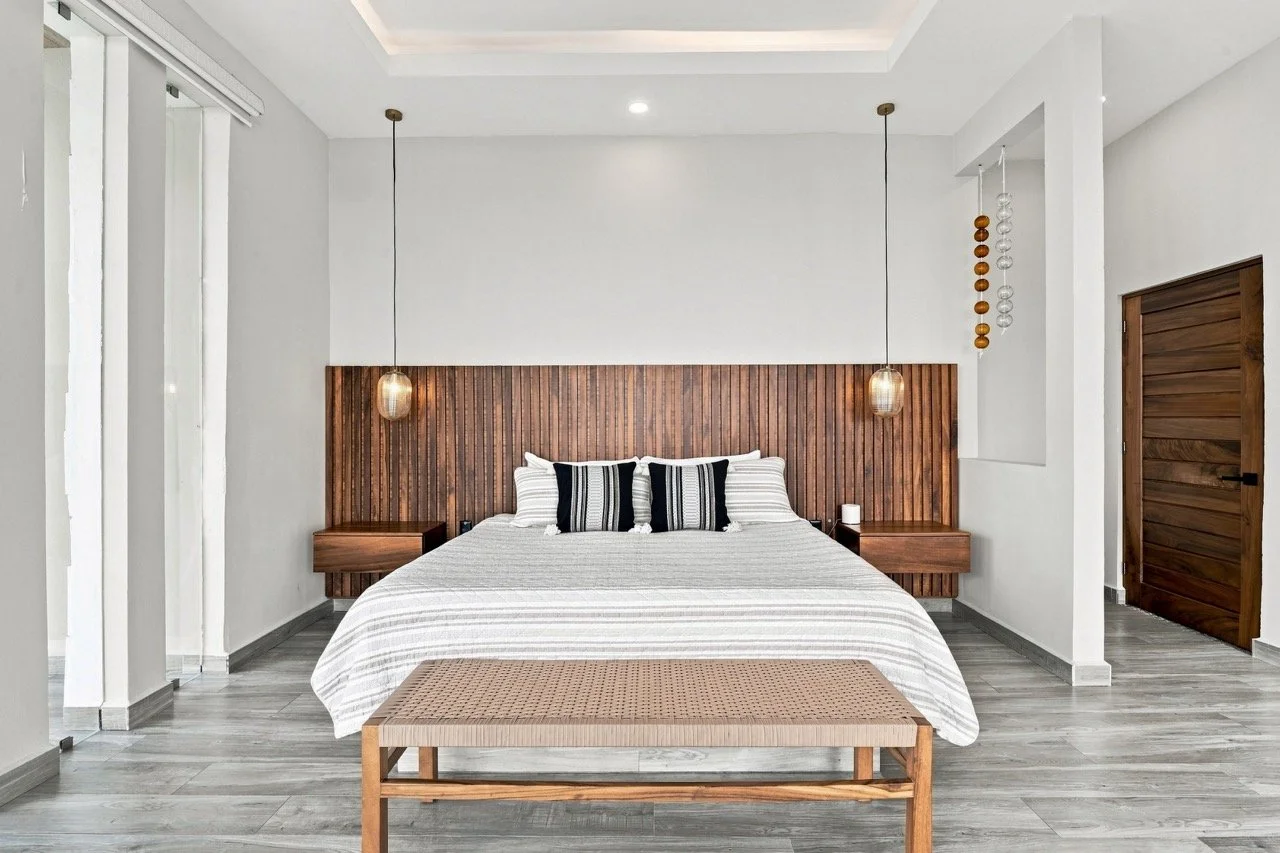 Habitación moderna con pared de madera en la cabecera, cama con sábanas blancas y cojines a rayas negras y blancas, lámparas colgantes a ambos lados, puerta de madera a la derecha y piso de madera clara.