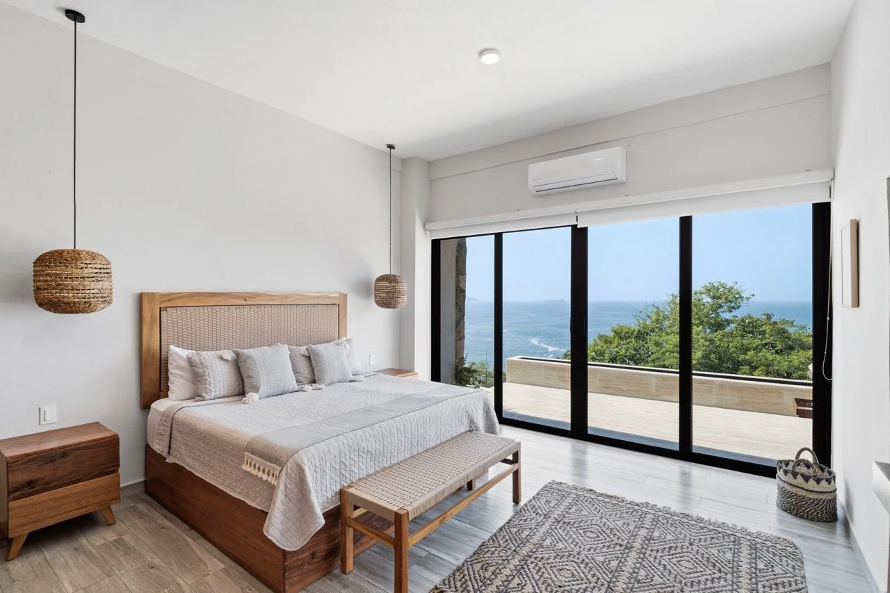 Habitación moderna con vista al mar, cama con ropa blanca y cojines, alfombra, mesas de madera, lámparas de tela, ventanas grandes con vista al océano y árboles.