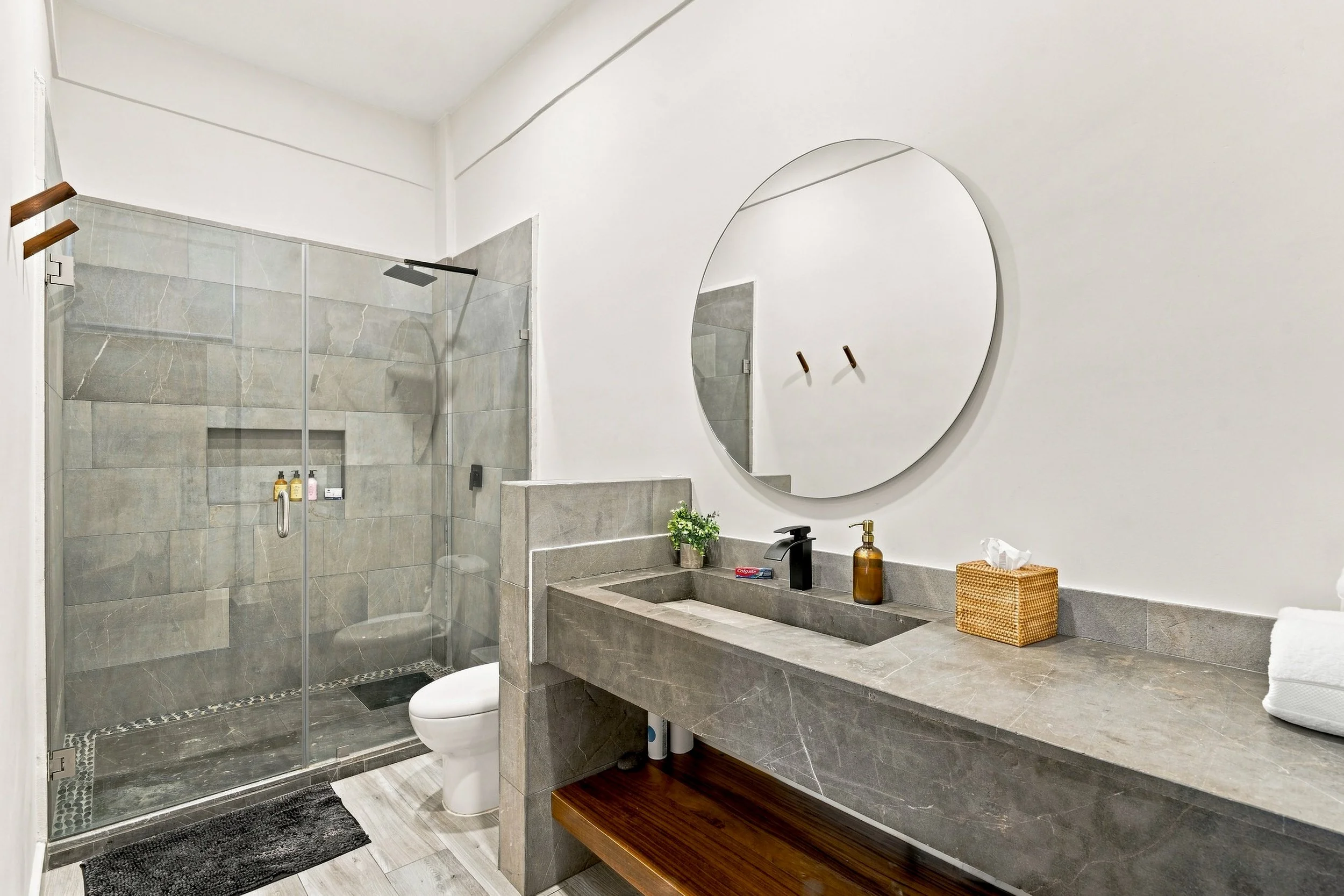 Baño moderno con ducha de vidrio, lavabo de piedra, espejo redondo y accesorios en tonos oscuros, con detalles decorativos y productos de higiene.