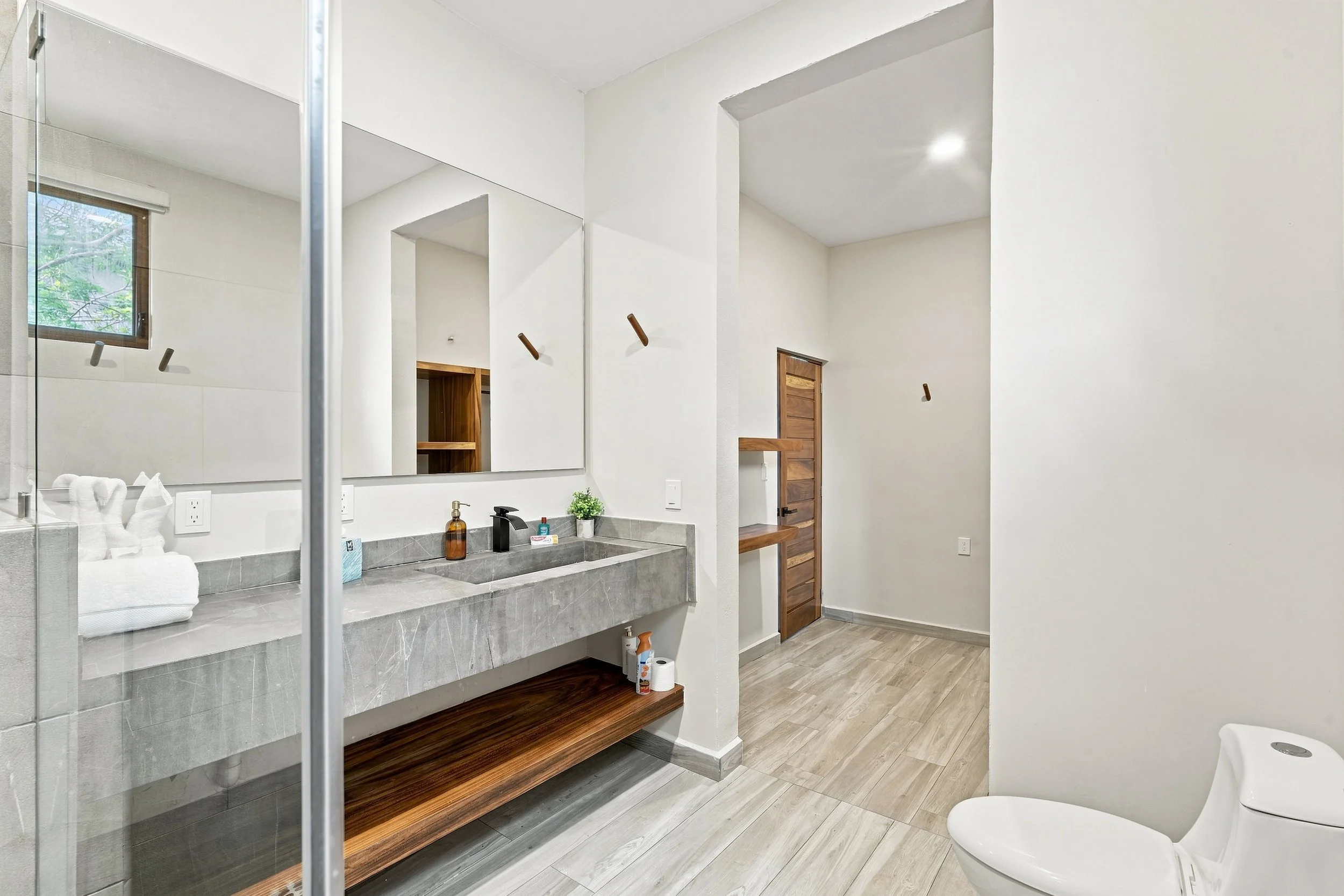 Baño moderno con lavabo de piedra, espejo, taza, papel higiénico, ventana, y ducha con puerta de vidrio.