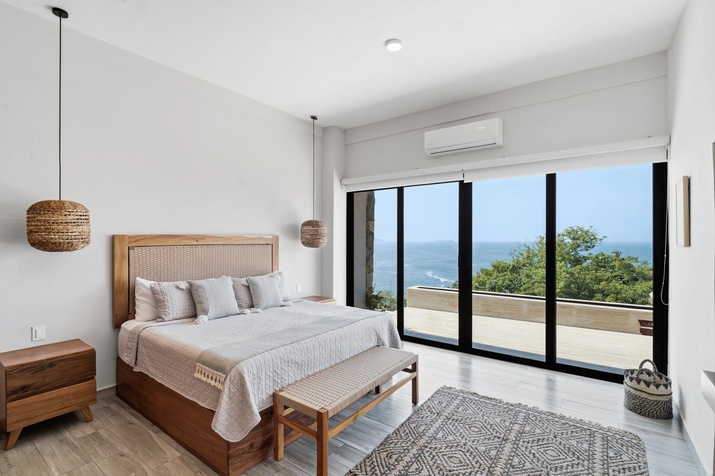Dormitorio con vista al mar, cama de madera, lámparas de mimbre, alfombra con patrón geométrico, y ventanas grandes con vista a la playa y árboles.
