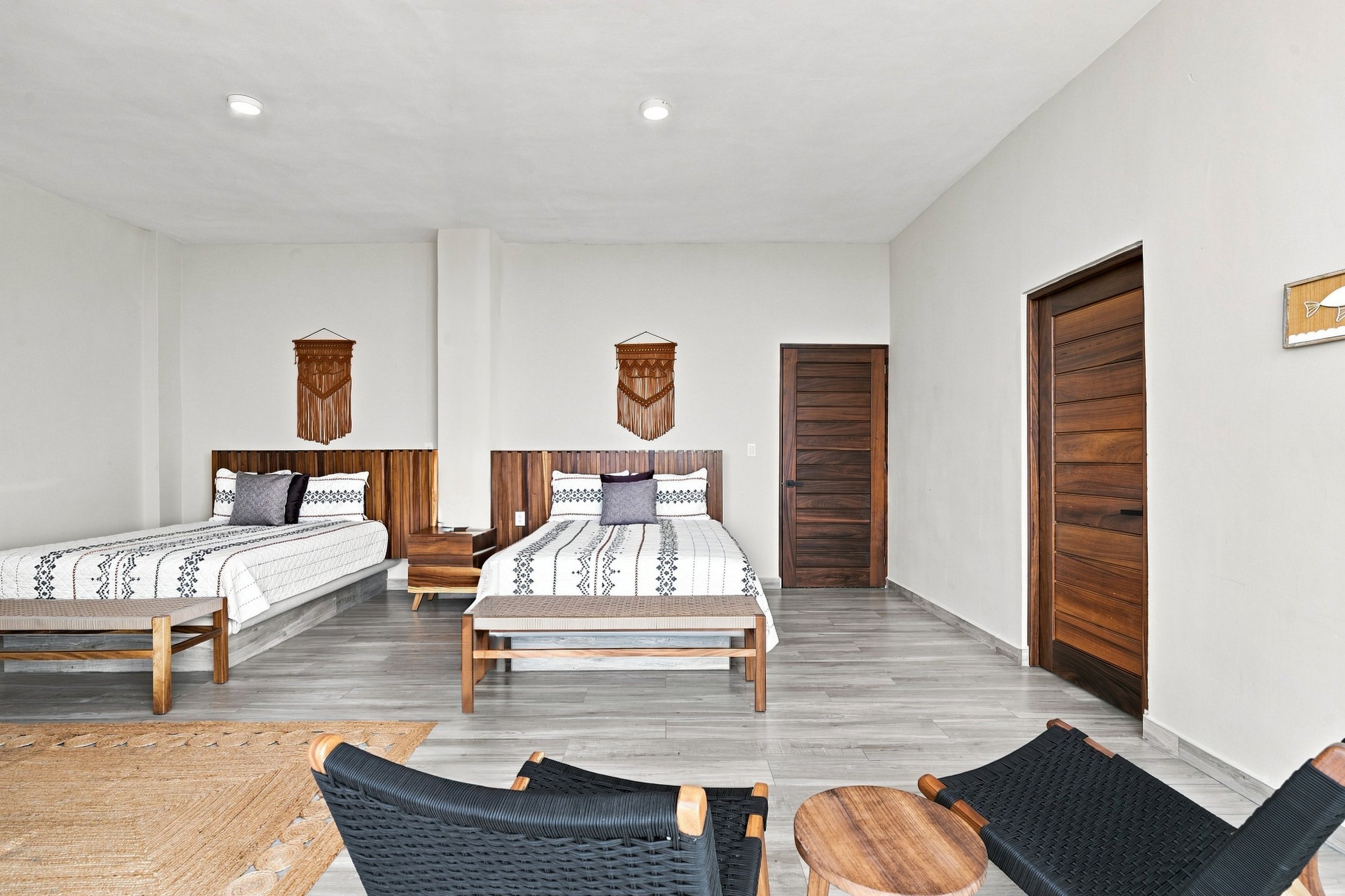 Habitación con dos camas con cabecera de madera, decoradas con mantas blancas con detalles en negro, y cojines grises y blancos. Paredes blancas, puertas de madera, y decoración en paredes con tapices de macramé. Muebles de madera y piso de laminado 