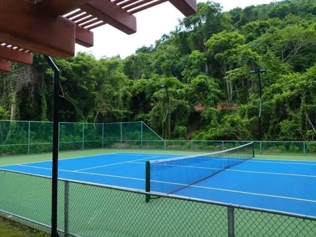 Cancha de tenis azul rodeada de una cerca, en un entorno de vegetación y árboles.