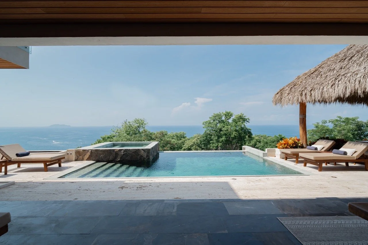 Vista de una piscina con jacuzzi en un deck de madera, con sillones y sombrillas de paja, vista al océano y al cielo despejado