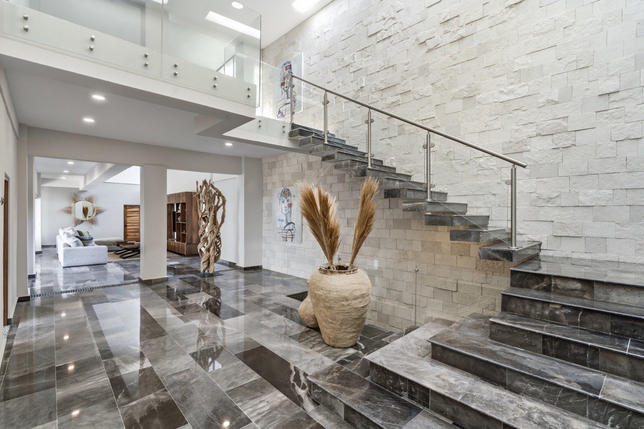Vestíbulo de una residencia moderna con pisos de mármol, una escalera de piedra con baranda de acero y una decoración minimalista, incluyendo un jarrón grande con ramas y obras de arte en la pared.