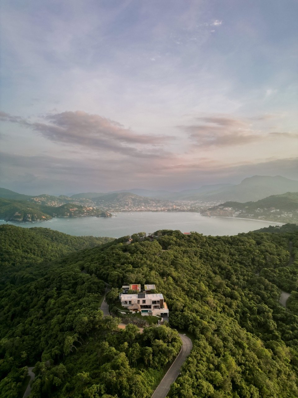 Vista aérea de una casa moderna en una colina rodeada de árboles verdes, con un lago y montañas al fondo, bajo un cielo parcialmente nublado.