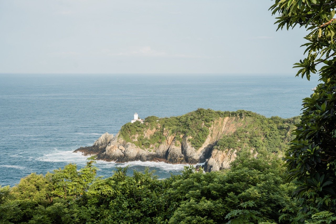 Vista de una isla con un faro en la orilla, rodeada de árboles verdes y el océano en el fondo