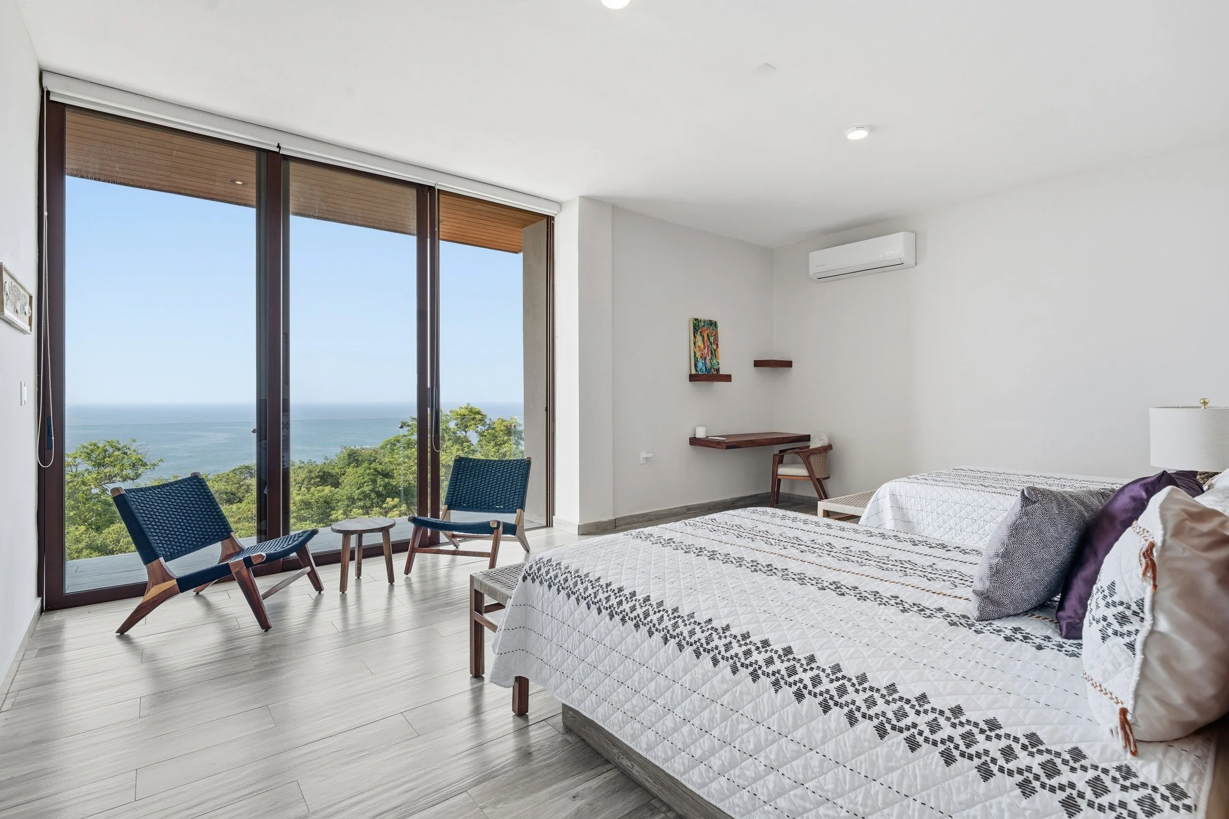 Habitación moderna con dos camas, vista a la naturaleza y al mar, ventanas grandes de vidrio, sillas de madera con asientos de malla, obra de arte en la pared, aire acondicionado.