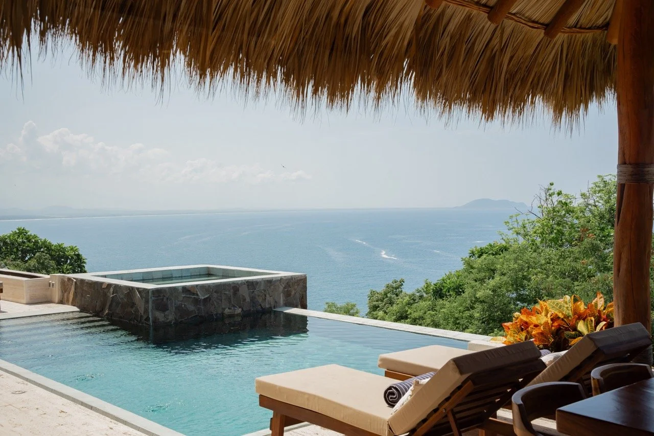 Vista de una piscina infinity con vista al mar desde una terraza con sombrilla de paja y tumbonas, rodeada de vegetación.